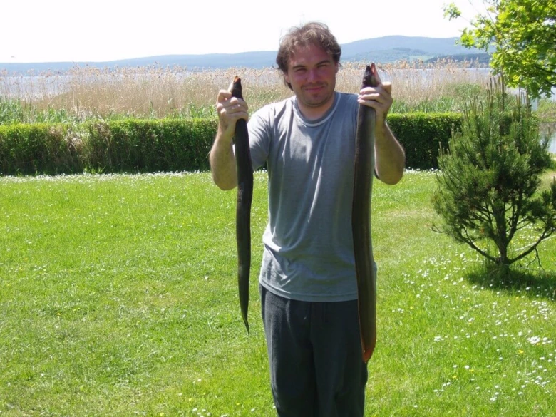 Muž drží dva úhoře v ruce na pozadí české krajiny.